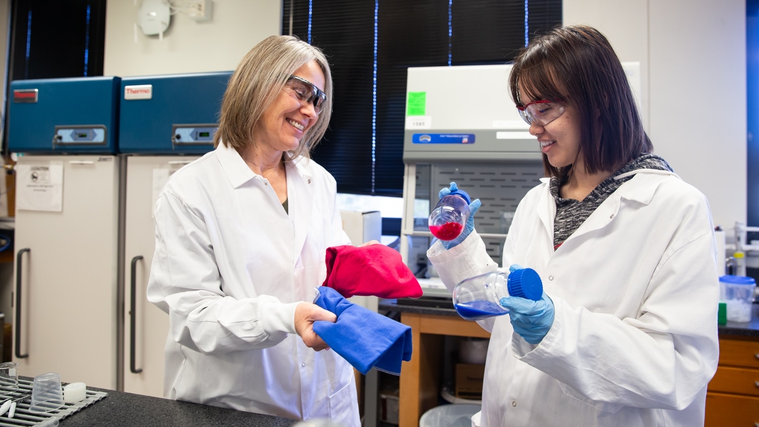 Sonja Salmon (left) and Siyan Wang (right) stand in the lab with lab coats and protective glasses. Salmon holds fabric samples and Wang holds two bottles of solution with some fabric fragments visible.