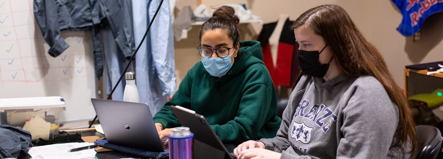 Two students work at a table in the Senior Design Lab. Textile samples are on the table they are working at, and the PPE suit they are working to improve is visible hanging on a whiteboard behind them.